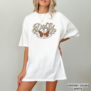 May include: A white short-sleeve t-shirt featuring a graphic print on the chest. The design includes the word Dolly in large cursive script, a central acoustic guitar with orange butterfly wings, and the text Tennessee Mountain Soul below.