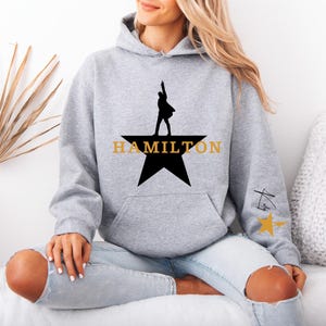 May include: A light gray hoodie featuring a black star with a silhouette of a person and the word "HAMILTON" in gold. The hoodie has a kangaroo pocket and a drawstring hood. A small design is on the sleeve.