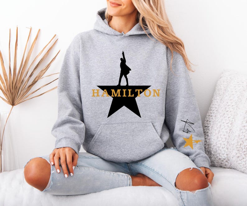 May include: A light gray hoodie featuring a black star with a silhouette of a person and the word "HAMILTON" in gold. The hoodie has a kangaroo pocket and a drawstring hood. A small design is on the sleeve.
