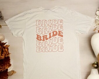 May include: A white t-shirt with the word "BRIDE" repeated five times in a retro, wavy font. The text is in a peachy-orange color.