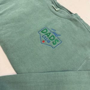May include: A seafoam green sweatshirt with the embroidered text "Cool Dads Club" in a blue and green design. The design includes a red star. The sweatshirt is made of a soft, comfortable material.