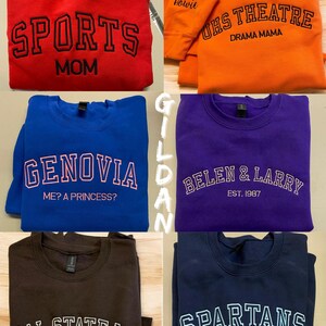 May include: A collection of crewneck sweatshirts in various colors, each featuring embroidered text. The sweatshirts include phrases like "SPORTS MOM," "UHS THEATRE DRAMA MAMA," "GENOVIA ME? A PRINCESS?," "BELEN & LARRY EST. 1987," "CAL STATE LA EST.1947," and "SPARTANS EST. 1953."
