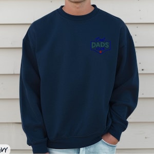 Puede incluir: Sudadera azul marino con el texto "Cool Dads Club" en un emblema azul, verde y rojo en el pecho. La sudadera es de cuello redondo y parece estar hecha de un material suave.