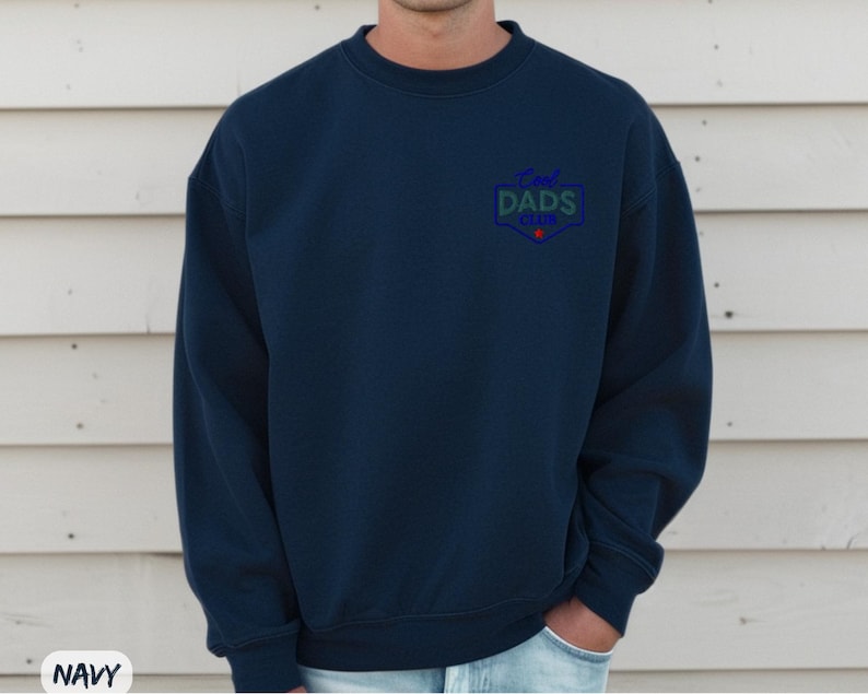 Puede incluir: Sudadera azul marino con el texto "Cool Dads Club" en un emblema azul, verde y rojo en el pecho. La sudadera es de cuello redondo y parece estar hecha de un material suave.