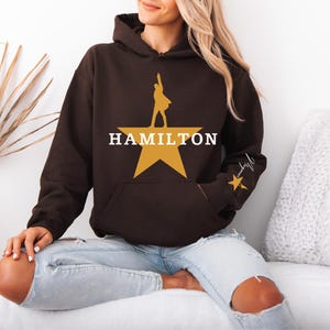 May include: A brown hoodie with a large gold star and the word "HAMILTON" in white. The hoodie features a silhouette of a person on the star and a smaller star with script on the sleeve. The hoodie has a front pocket and a hood.