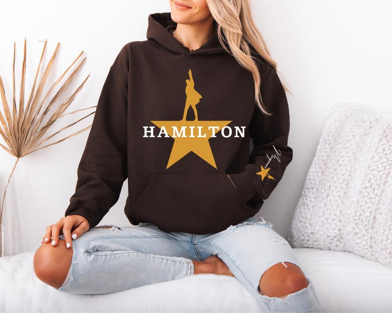 May include: A brown hoodie with a large gold star and the word "HAMILTON" in white. The hoodie features a silhouette of a person on the star and a smaller star with script on the sleeve. The hoodie has a front pocket and a hood.