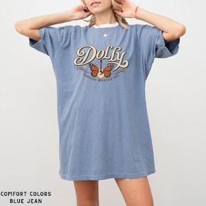 May include: A blue jean colored short-sleeve t-shirt featuring a graphic print with the text Dolly and Tennessee Mountain Soul. The design includes a central acoustic guitar with orange butterfly wings and small white stars on a cream background.