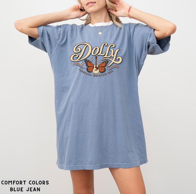 May include: A blue jean colored short-sleeve t-shirt featuring a graphic print with the text Dolly and Tennessee Mountain Soul. The design includes a central acoustic guitar with orange butterfly wings and small white stars on a cream background.