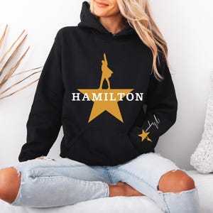 May include: Black hooded sweatshirt featuring a gold star design with a silhouette and the word "HAMILTON" in white. The sleeve has a signature and a small gold star. The sweatshirt has a front pocket.