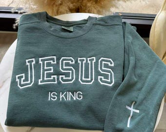 May include: A dark green sweatshirt with the words "JESUS IS KING" in white block letters. A white cross is on the left sleeve. The sweatshirt is folded on a white marble-topped table.