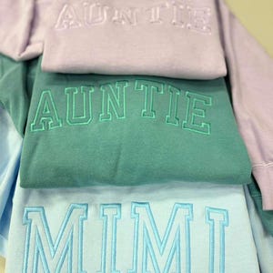 May include: A stack of three sweatshirts in pastel colors: lavender, teal, and light blue. Each sweatshirt has embroidered text: "AUNTIE" on the lavender and teal sweatshirts, and "MIMI" on the light blue sweatshirt. The sweatshirts are folded.