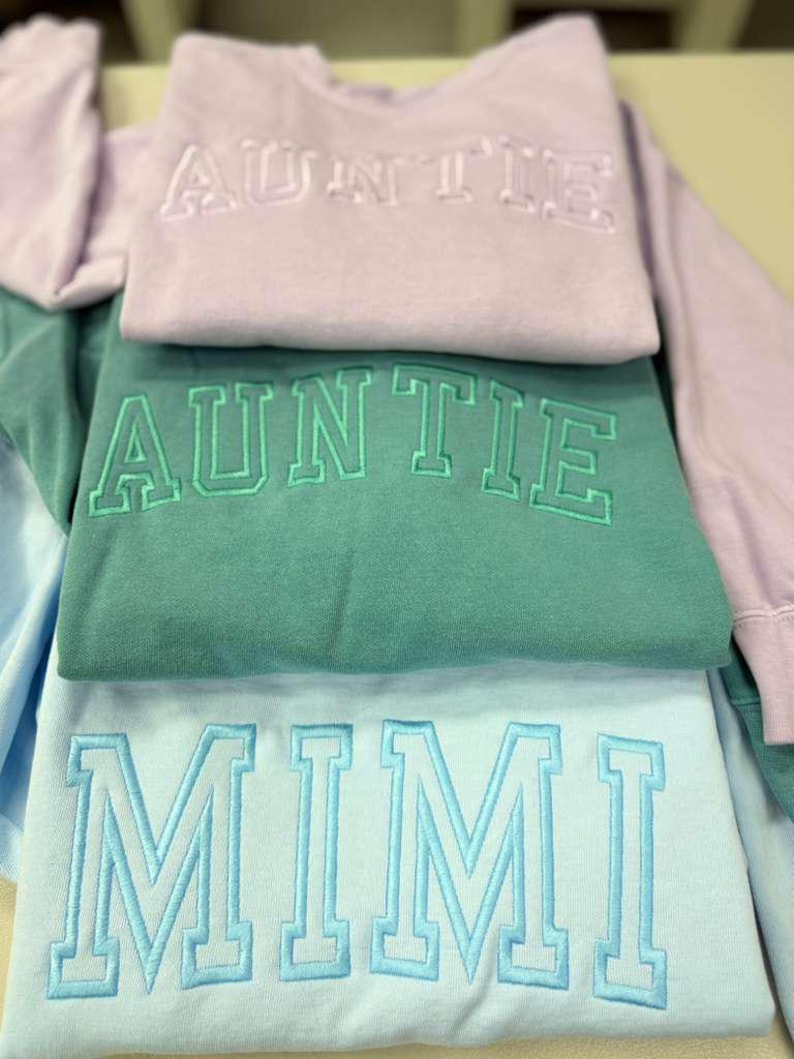 May include: A stack of three sweatshirts in pastel colors: lavender, teal, and light blue. Each sweatshirt has embroidered text: "AUNTIE" on the lavender and teal sweatshirts, and "MIMI" on the light blue sweatshirt. The sweatshirts are folded.