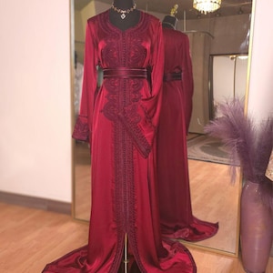 May include: A long, flowing burgundy dress with intricate embroidery and a matching belt. The dress features long sleeves and a high neckline. The dress is displayed on a mannequin in front of a mirror.