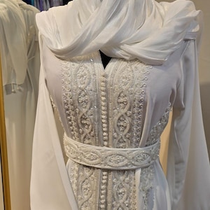 May include: A white dress with intricate embroidery and a matching white scarf. The dress has a long, flowing silhouette and a cinched waist with a beaded belt.
