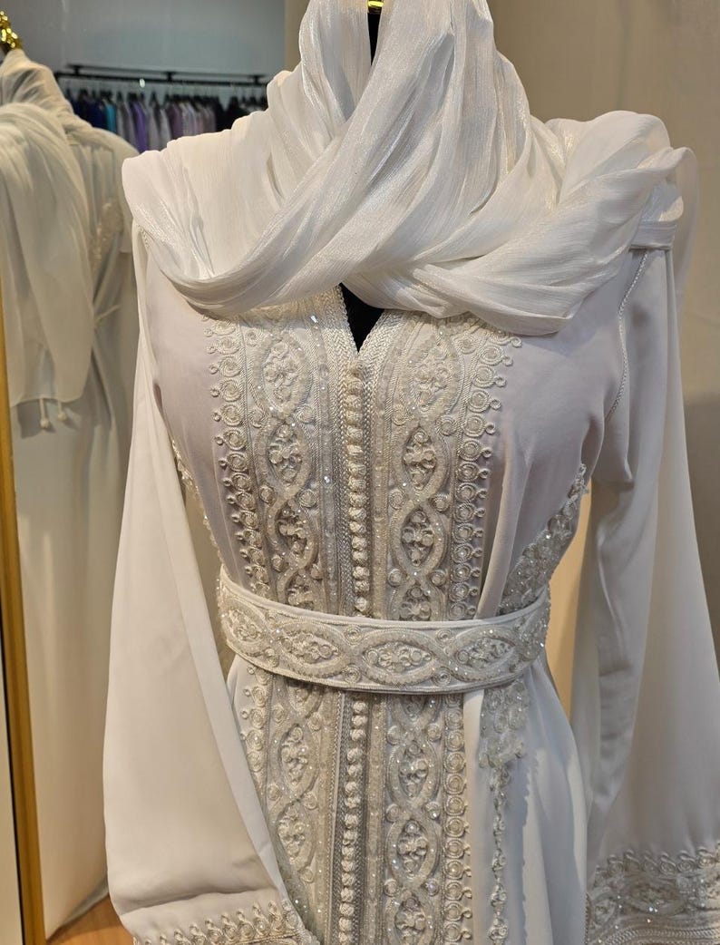 May include: A white dress with intricate embroidery and a matching white scarf. The dress has a long, flowing silhouette and a cinched waist with a beaded belt.