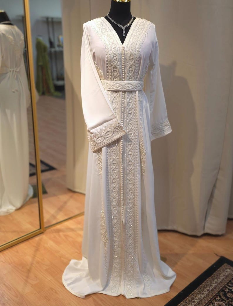May include: A white kaftan with intricate embroidery and pearl embellishments. The kaftan has a V-neckline, long sleeves, and a belt at the waist.