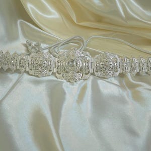 以下が含まれることがあります： 繊細なフィリグリー模様が施されたシルバーカラーのベルト。中央に正方形のバックルがあり、両サイドのパネルは繊細なチェーンで繋がれています。クリーム色のサテン生地の上に展示されています。