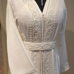 May include: A white kaftan with intricate embroidery and beading. The kaftan has a V-neckline and long sleeves. It is belted at the waist.