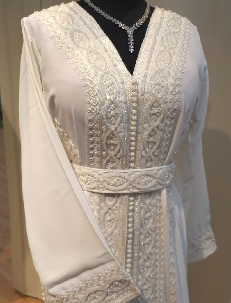 May include: A white kaftan with intricate embroidery and beading. The kaftan has a V-neckline and long sleeves. It is belted at the waist.