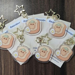 May include: Six clear acrylic keychains featuring a cartoon rabbit character sitting in a slice of bread with a fried egg. The keychains are attached to gold star-shaped keyrings. The text "Rabbitkero" is visible on one of the keychains.