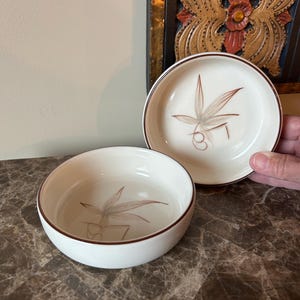 May include: Two white ceramic bowls with a brown rim and a stylized plant design in the center. One bowl is held by a hand. The bowls are on a marble surface.