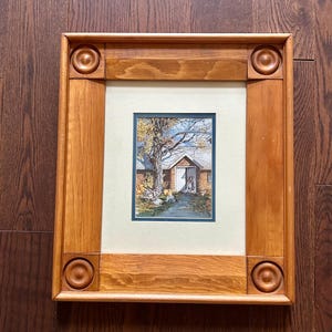 May include: A framed watercolor painting of a house with an open door, a tree, and a path. The frame is made of light brown wood with decorative circles at each corner. The painting is set against a light green mat.