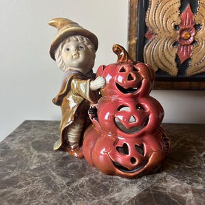 May include: A ceramic figurine of a child wearing a pointed hat and a brown cloak, standing beside a stack of three carved, glossy red-orange pumpkins with cut-out facial features. The pumpkins are arranged vertically to form a hollow structure.