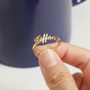 Puede incluir: Un anillo dorado con el nombre "Tiffany" escrito en cursiva.