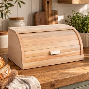 May include: A light-colored wooden bread box featuring a roll-top lid with a small rectangular handle. The bread box has a curved front design constructed from horizontal wooden slats and a solid base, providing a storage solution.