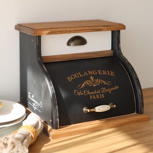 May include: A black and white wooden bread box with a roll-top door featuring gold text that reads BOULANGERIE Cafe Chaud et Beignets PARIS. The unit includes a top drawer with a bronze handle and a white ceramic handle on the main door.