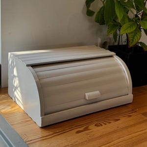 May include: A white, wooden bread box with a roll-top lid and a small handle. The box is rectangular with rounded edges and sits on a wooden surface. A plant in a black pot is visible in the background.