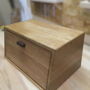 May include: A wooden bread box with a light brown finish. The box has a rectangular shape with a curved top and a small, dark brown handle. The wood grain is visible on the surface.