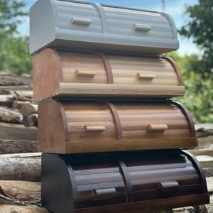 Op de afbeelding: Vier houten broodtrommels op elkaar gestapeld. De bovenste is lichtgrijs geschilderd, de volgende twee zijn van natuurlijk hout en de onderste is donkerbruin. Elk heeft een gebogen bovenkant en een klein handvat. De broodtrommels zijn van hout.