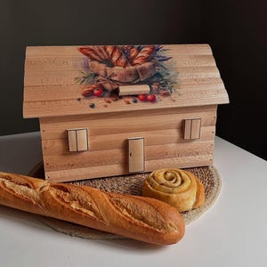 May include: A wooden bread box shaped like a house, with a painted illustration of bread and berries on the roof. A baguette and a cinnamon roll sit in front of the box on a woven mat.