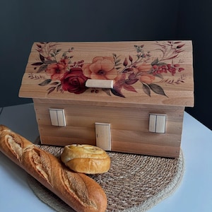 May include: Wooden bread bin shaped like a house, with a floral design on the roof. The bread bin has a door and two windows. A loaf of bread and a roll sit on a woven mat in front of the bin.