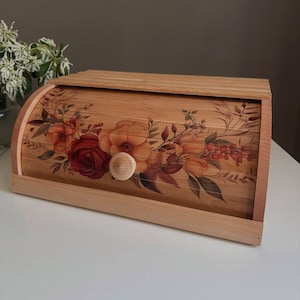Op de afbeelding: Houten broodtrommel met een roldeksel. De voorkant is versierd met een bloemenontwerp met rode, oranje en bruine bloemen. Het heeft een houten knop. De broodtrommel staat op een wit oppervlak.