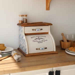Puede incluir: Una panera de madera con puerta enrollable color crema y un cajón superior. El frente presenta texto negro que dice BOULANGERIE Cafe Chaud et Beignets PARIS. Los lados tienen un patrón floral azul y blanco, y los tiradores son de cerámica.