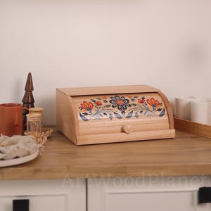 Puede incluir: Panera de madera clara con tapa enrollable. La panera presenta un diseño floral en naranja, azul y blanco. Un pequeño pomo de madera está en la parte delantera. La panera está sobre una encimera de madera.