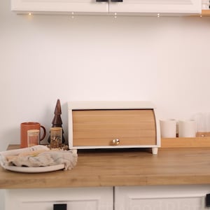May include: A white and wood breadbox with a roll-top door sits on a wooden countertop. The kitchen scene includes white cabinets, a mug, and decorative items. The breadbox has a gold knob.