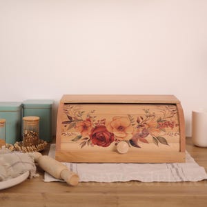 May include: Wooden bread box with a floral design featuring red, orange, and yellow roses. The box has a curved lid and a small wooden knob. The bread box is sitting on a white and gray striped cloth.