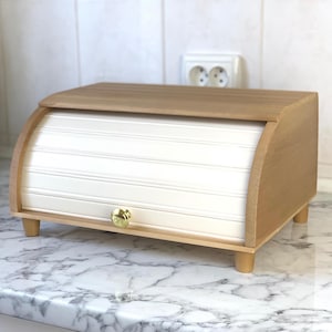 Puede incluir: Panera de madera con puerta enrollable y pomo de latón. La panera es de madera clara y tiene una parte superior curvada. La puerta delantera es blanca con ranuras horizontales. La panera se apoya en cuatro patas pequeñas de madera.