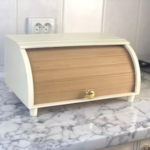 Op de afbeelding: Een witte en natuurlijke houten broodtrommel met een roldeur en een goudkleurige knop. De broodtrommel heeft een gebogen ontwerp en staat op kleine, witte voetjes. De bovenkant is ook wit met horizontale groeven.