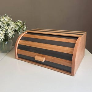 Puede incluir: Panera de madera con tapa enrollable. La panera presenta franjas alternas de madera clara y oscura. Un pequeño tirador de madera está en la parte delantera. La panera está sobre una superficie blanca.
