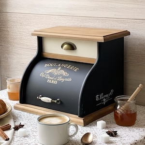 May include: A black wooden bread box with a natural wood top and base, featuring a cream-colored drawer with a brass handle. The front panel displays the gold text BOULANGERIE, Grains, Céréales et Levains, and PARIS, with a white ceramic handle.