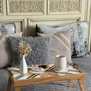 Puede incluir: Una bandeja de desayuno de madera con un patrón geométrico en blanco, gris y marrón. La bandeja tiene patas plegables y está colocada sobre una manta gris y peluda. Una taza blanca con un patrón blanco y gris está en la bandeja, junto con un plato de pequeños dulces.