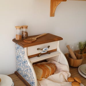 Puede incluir: Una panera de madera con un patr&oacute;n floral azul y blanco en los laterales, que cuenta con un caj&oacute;n con dos asas de metal ornamentadas. La abertura frontal muestra pan rebanado sobre un pa&ntilde;o claro, con dos frascos de vidrio encima.