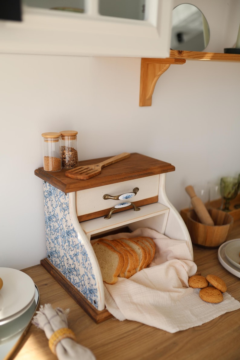 Puede incluir: Una panera de madera con un patr&oacute;n floral azul y blanco en los laterales, que cuenta con un caj&oacute;n con dos asas de metal ornamentadas. La abertura frontal muestra pan rebanado sobre un pa&ntilde;o claro, con dos frascos de vidrio encima.
