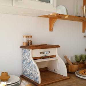 Puede incluir: Una panera de madera con un caj&oacute;n blanco y paneles laterales con un patr&oacute;n floral azul y blanco. La unidad incluye un caj&oacute;n extra&iacute;ble con un tirador decorativo y un compartimento de almacenamiento abierto en la parte inferior con base de madera.