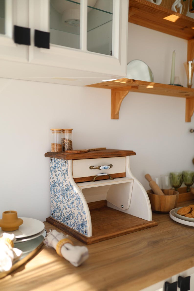Puede incluir: Una panera de madera con un caj&oacute;n blanco y paneles laterales con un patr&oacute;n floral azul y blanco. La unidad incluye un caj&oacute;n extra&iacute;ble con un tirador decorativo y un compartimento de almacenamiento abierto en la parte inferior con base de madera.