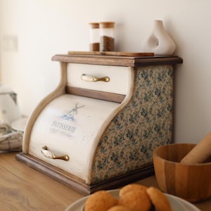Pode incluir: Uma caixa de pão de madeira com porta de enrolar com um gráfico de moinho de vento azul e o texto PATISSERIE DE PARIS. As laterais têm um padrão floral azul e a unidade inclui uma gaveta superior com puxador dourado e um puxador a condizer.